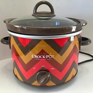 May include: A Crock-Pot slow cooker with a glass lid and brown handle. The exterior features a red, brown, and gold chevron pattern. The control knob has settings for Off, Low, and High.
