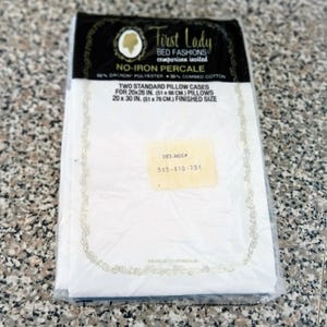 May include: A package of two white First Lady brand standard pillowcases. The package reads "No-Iron Percale" and lists the dimensions as 20x26 inches (51 x 66 cm) and 20 x 30 inches (51 x 76 cm) finished size. The pillowcases are made of 65% Dacron polyester and 35% combed cotton.