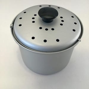 May include: A silver-colored metal cooking pot with a matching lid. The lid has several small holes and a black knob. The pot has a handle on each side. The pot is designed for cooking.