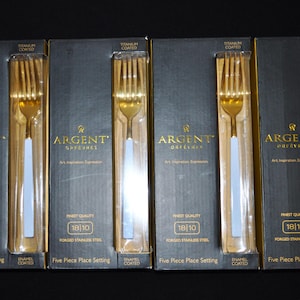 Puede incluir: Cinco cajas de cubiertos de color dorado con mangos blancos. Cada caja tiene el texto "Argent Orfevres" y "Finest Quality 18/10 Forged Stainless Steel" en la parte delantera. Las cajas están etiquetadas como "Titanium Coated" y cada caja contiene un juego de cubiertos de cinco piezas.