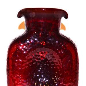May include: A deep red glass vase with a textured, dimpled surface and a wide, flared rim. The vessel features a rounded, square-like body with a translucent quality that allows light to pass through, highlighting the rich crimson color throughout.
