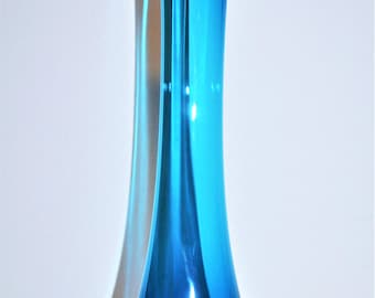 Pode incluir: Um vaso de vidro azul, alto e elegante, com a parte superior alargada. O vaso tem uma forma esguia e alongada e uma base azul escuro que transita para um azul mais claro e vibrante em direção ao topo. O vaso tem cerca de 30 cm de altura.