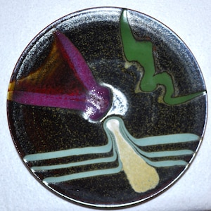 May include: A black ceramic plate with abstract designs in shades of green, purple, yellow, and brown. The plate has a speckled glaze.