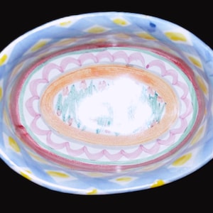 May include: Hand-painted ceramic oval bowl with a colorful design. The rim features a blue and white pattern with yellow accents. The interior has a red border, pink scalloped design, and an orange and green center.