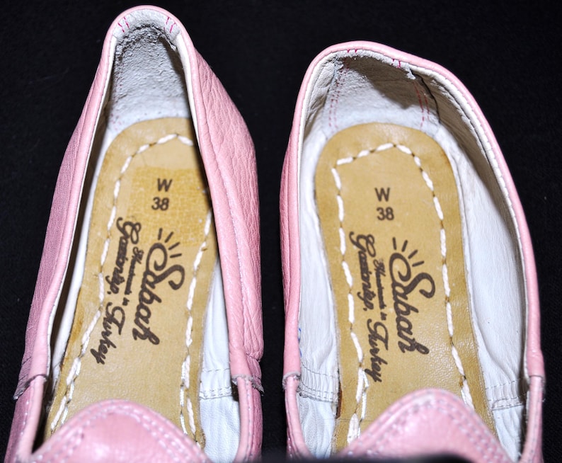 May include: A pair of pink leather ballet flats with a white leather lining. The inside of the shoes has a label that reads "Sıbaf Handmade in Turkey" and "W 38".