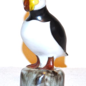 May include: A ceramic figurine of a puffin standing on a cylindrical base. The bird features a white chest, black back and head, and a distinctive beak coloured in yellow, red, and brown. The base has a mottled grey and brown glazed finish.