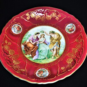 May include: A red and gold porcelain plate with a raised rim and a central image of three figures in a garden setting. The figures are painted in shades of blue, green, and brown. The plate is decorated with gold leaf and floral motifs.
