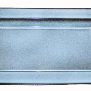 Puede incluir: Una fuente rectangular de cerámica azul claro con una superficie ligeramente texturizada. La fuente tiene un borde elevado y angulado y un acabado brillante, adecuada para servir aperitivos o platos pequeños.