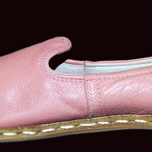 May include: A pair of pink leather slip-on shoes with a white stitched trim around the sole.