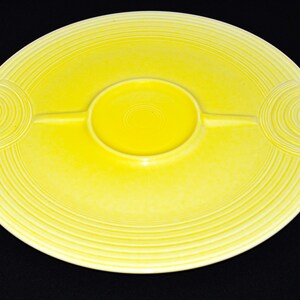 Pode incluir: Um prato de servir de cerâmica amarelo com uma tigela central para molhos e dois compartimentos laterais. O prato tem um design de círculos concêntricos.