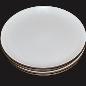 May include: A stack of three round ceramic plates with a smooth, matte white surface on the top and a contrasting brown, unglazed rim around the edge of each plate, viewed from a high angle against a solid black background.