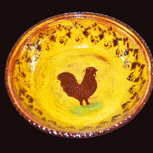Op de afbeelding: Een handgemaakte keramische kom met een gele binnenkant en een bruin hanenontwerp. De rand is bruin met een gespikkeld patroon. De haan is bruin met een gestippelde textuur, staand op een groene vlek. De kom wordt waarschijnlijk gebruikt voor het serveren van voedsel.