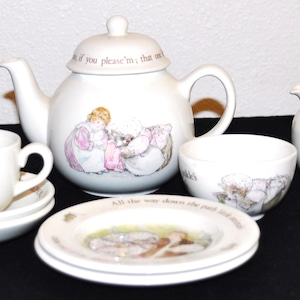 Peut inclure: Un service à thé en céramique blanche orné d'illustrations de Mrs. Tiggy-winkle. L'ensemble comprend une théière avec couvercle, un pot à lait, une tasse avec soucoupe et un petit bol. Chaque pièce présente du texte imprimé et des motifs colorés.