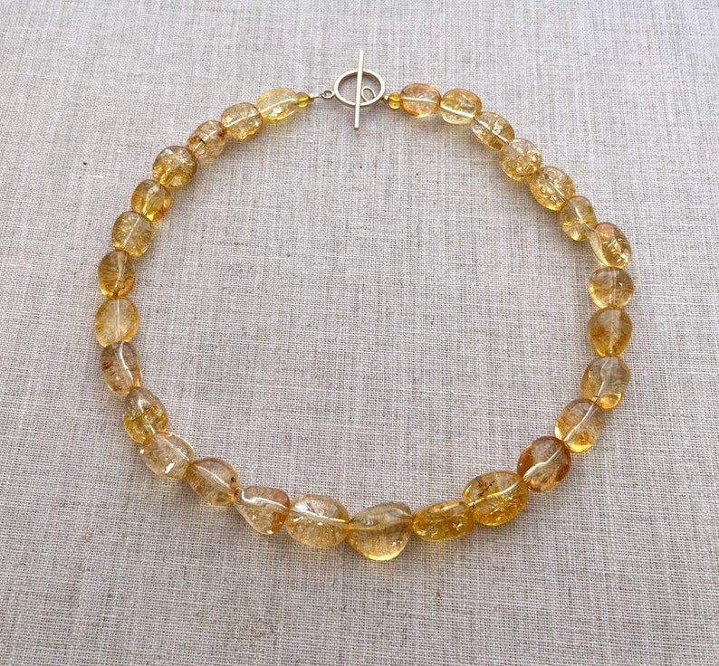 May include: A gold necklace made of oval-shaped, translucent, yellow citrine beads. The necklace has a silver toggle clasp.