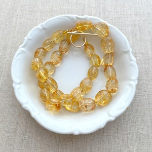 May include: A strand of oval-shaped, translucent, golden citrine beads with a gold clasp. The beads are arranged in a circle on a white plate.