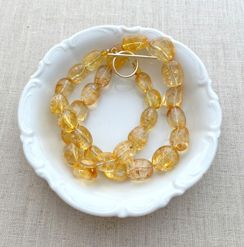 May include: A strand of oval-shaped, translucent, golden citrine beads with a gold clasp. The beads are arranged in a circle on a white plate.