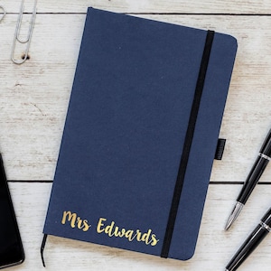 May include: A navy blue notebook with a black elastic closure and gold lettering that reads "Mrs Edwards".