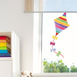 Op de afbeelding: Een kleurrijke regenboogvlieger met een groene staart en paarse, blauwe en roze strikken zit op een raam in een kinderkamer. De vlieger is een raamsticker.