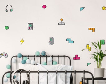 Op de afbeelding: Een zwart metalen bedframe met een wit-blauw gestreept dekbed. De muur achter het bed is versierd met kleurrijke stickers van videogamecontrollers, spelfiguren en gepixelde blokken.