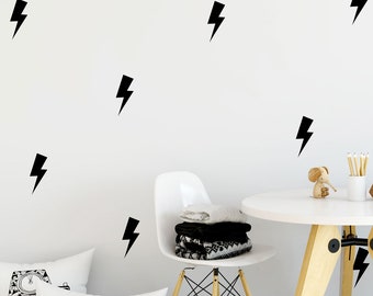 May include: A white wall with black lightning bolt decals. A white chair with a black and white blanket on it. A white table with a wooden base. A wooden elephant figurine and a glass vase with pencils on the table. A gray and white polka dot rug on the floor. Two white pillows with black and white camera designs.
