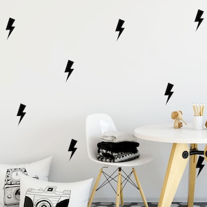 May include: A white wall with black lightning bolt decals. A white chair with a black and white blanket on it. A white table with a wooden base. A wooden elephant figurine and a glass vase with pencils on the table. A grey and white polka dot rug on the floor. Two white pillows with black and white camera designs.