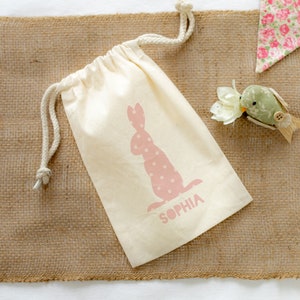 Puede incluir: Una bolsa de algodón blanco con cordón de ajuste, adornada con una silueta de conejo rosa y el nombre "Sophia" impreso. La bolsa está sobre una superficie de arpillera marrón.