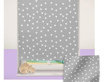 May include: Gray blackout roller shade with a white heart pattern. The shade is fully extended and shows a view of a beach through a window. A sample of the fabric is shown below the window.