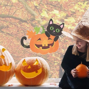 May include: A young girl wearing a witch's hat and a black dress holds a small orange pumpkin. She is standing in front of a window with a fall foliage background. There are two carved pumpkins and a black cat holding a pumpkin on the window.