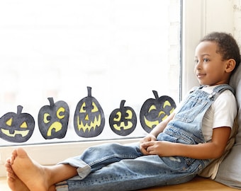 May include: A young person wearing blue overalls sits by a window with black and yellow pumpkin cutouts on the glass. The person is looking out the window.