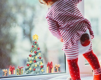 May include: A young child in a red and white striped outfit looks out a window at Christmas window clings. The window clings include a Christmas tree, presents, and a star.