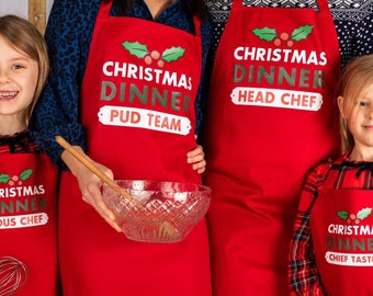 Puede incluir: Cuatro delantales rojos con texto festivo de temática de cena de Navidad. Los delantales tienen acebo verde y bayas rojas. El texto en los delantales dice "Christmas Dinner Sous Chef", "Christmas Dinner Pud Team", "Christmas Dinner Head Chef" y "Christmas Dinner Chief Taster".
