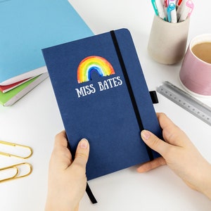 Puede incluir: Una libreta azul marino con un arco&iacute;ris y el texto "MISS BATES" en la portada. La libreta est&aacute; siendo sostenida por las manos de una persona.