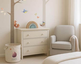 May include: A nursery scene with a cream-colored dresser, a gray armchair, and a round storage bin. The wall features tree decals, birds, and a wooden rainbow. The storage bin has a hedgehog graphic and the name "GRACE".