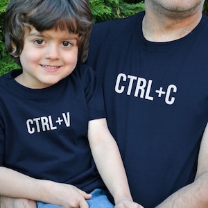 Könnte beinhalten: Ein Vater und sein Sohn tragen passende marineblaue T-Shirts. Das Shirt des Vaters sagt "CTRL+C" und das Shirt des Sohnes sagt "CTRL+V".