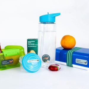 May include: A green Sistema lunchbox with a blue and green label that says "Adam Williams". A clear plastic water bottle with a blue lid and a straw. A small blue container with a blue and green label that says "Adam Williams". A blue lunchbox with a green and blue label that says "Adam Williams". A small orange fruit and a red sweet.