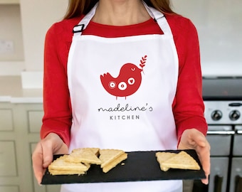 Puede incluir: Delantal blanco con un diseño de pájaro rojo y el texto "madeline's KITCHEN". Una persona sostiene una bandeja de pizarra negra con galletas de mantequilla.