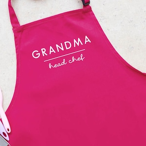 May include: Pink kitchen apron with white text that reads "Grandma head chef".