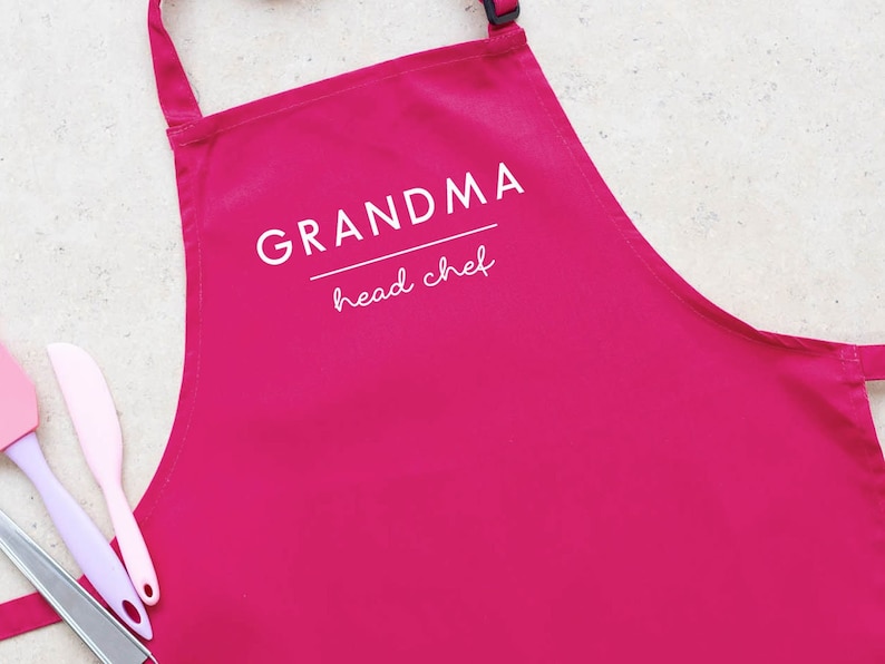 May include: Pink kitchen apron with white text that reads "Grandma head chef".