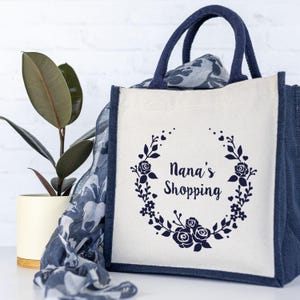 May include: A white canvas tote bag with navy blue trim and handles. The bag has a floral wreath design with the text "Nana's Shopping" in the center.