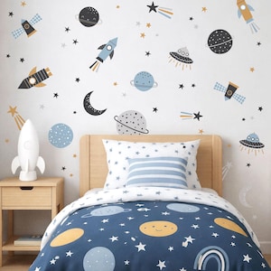 May include: A bedroom with a space theme, featuring a bed with a blue duvet cover adorned with planets and stars. The white walls are decorated with rockets, planets, stars, and a crescent moon. A white rocket lamp sits on a wooden bedside table.