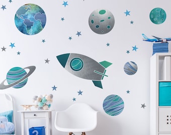 May include: A set of space-themed wall decals featuring planets, stars, and a rocket ship. The planets are painted in shades of blue and green, while the stars are silver. The rocket ship is silver with a green tail.