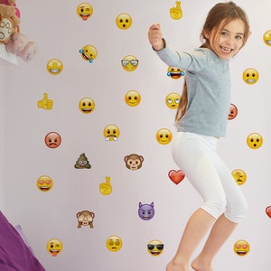 May include: A young girl jumps in front of a wall covered in yellow emoji stickers. The stickers include a variety of expressions, such as winking, smiling, and crying.