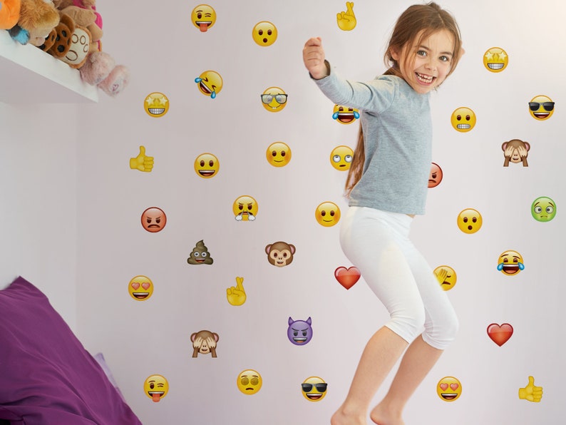May include: A young girl jumps in front of a wall covered in yellow emoji stickers. The stickers include a variety of expressions, such as winking, smiling, and crying.