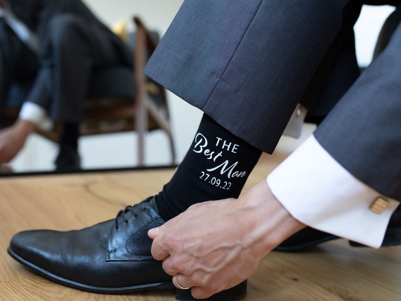 Peut inclure: Un homme met une chaussette noire avec du texte blanc qui dit "The Best Man 27.09.22". Il porte un costume noir et des chaussures habill&eacute;es noires.