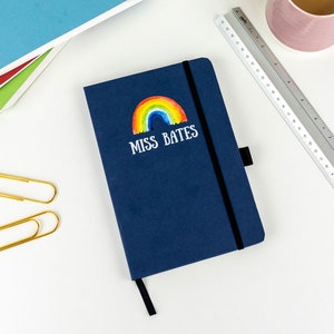 Puede incluir: Un cuaderno azul oscuro con un arco&iacute;ris y el texto "Miss Bates" en la portada. El cuaderno est&aacute; sobre una superficie blanca con una regla, dos clips de papel dorados y una pila de cuadernos de colores.