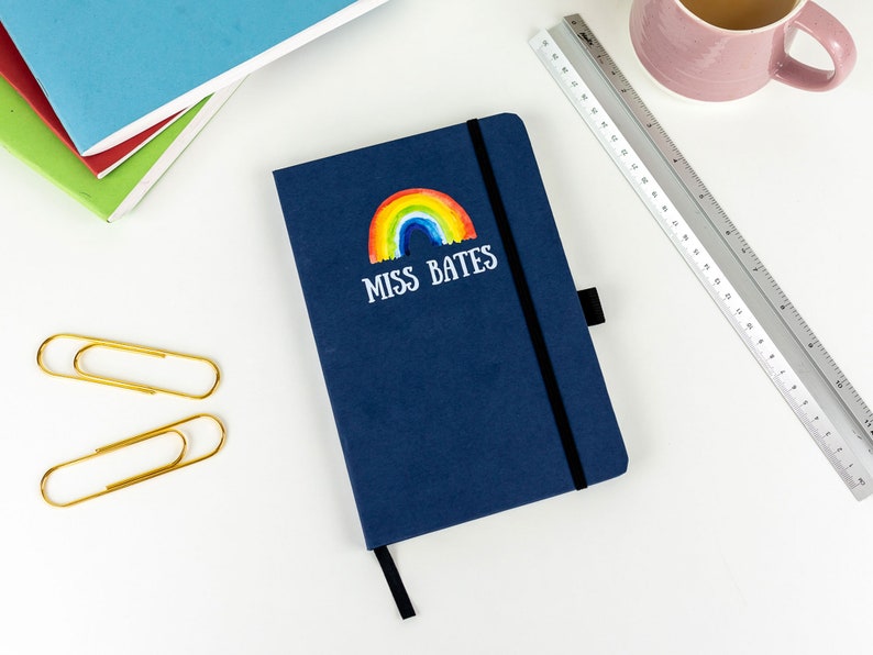 Puede incluir: Un cuaderno azul oscuro con un arco&iacute;ris y el texto "Miss Bates" en la portada. El cuaderno est&aacute; sobre una superficie blanca con una regla, dos clips de papel dorados y una pila de cuadernos de colores.