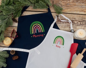 Puede incluir: Dos delantales navideños con un diseño de arcoíris. El delantal azul tiene la palabra "Mummy" bordada, y el delantal blanco tiene la palabra "Riley" bordada.