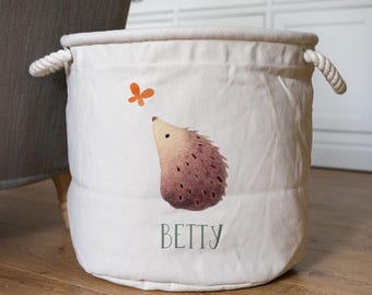 May include: A white canvas storage bin with rope handles. The bin features a watercolor illustration of a hedgehog and an orange butterfly. The name "BETTY" is printed in green script below the hedgehog. This bin is designed for storing toys or other items.