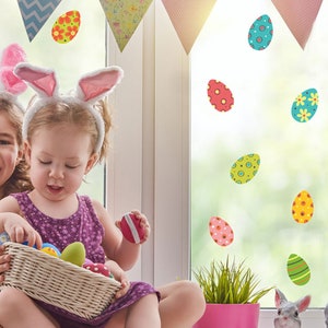 May include: Two young girls wearing bunny ears are holding a basket of colourful Easter eggs. The window behind them is decorated with colourful Easter egg stickers.