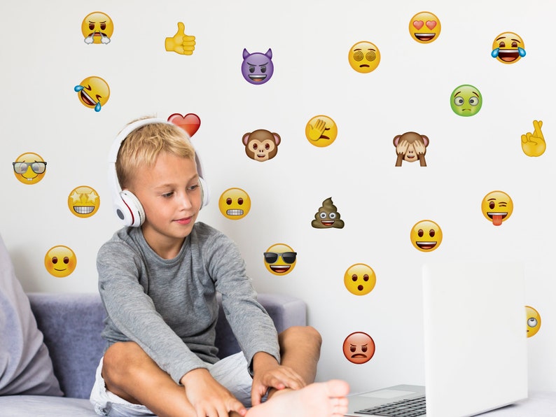 May include: A young boy wearing headphones sits on a white couch and looks at a laptop computer. The wall behind him is decorated with a variety of emoji stickers, including a thumbs up, a heart, a monkey, a poop emoji, and a winking face.